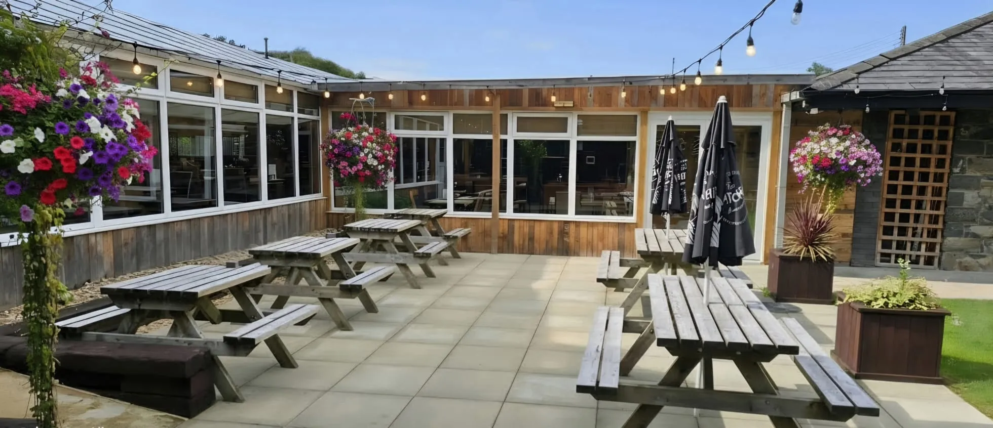 The terrace beside the pub: picnic benches on the paving, hanging baskets and festoon lights above