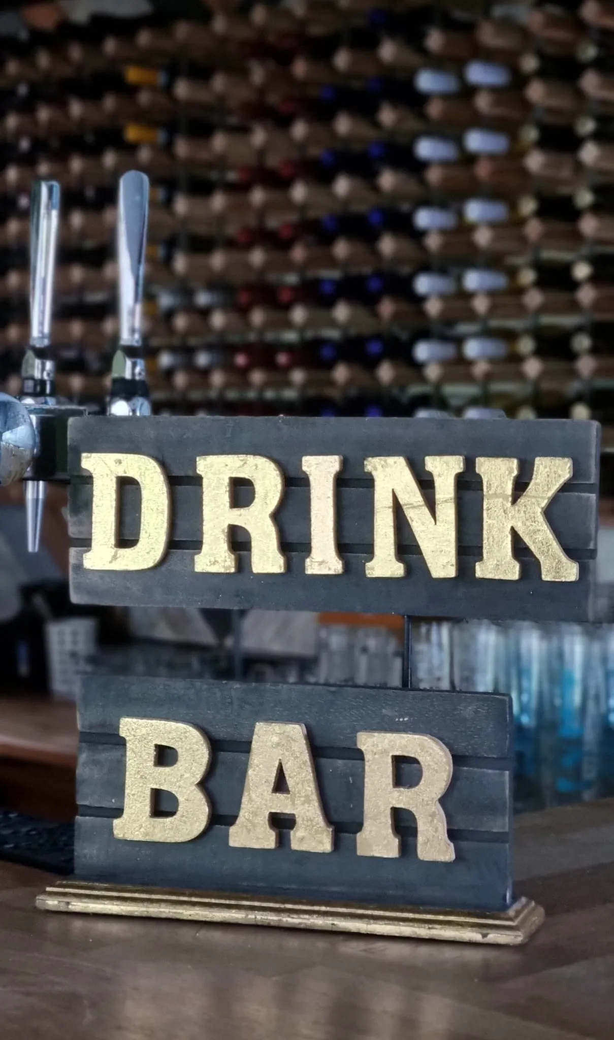 A gold drink sign on the bar top with a wall of corked wine bottles behind