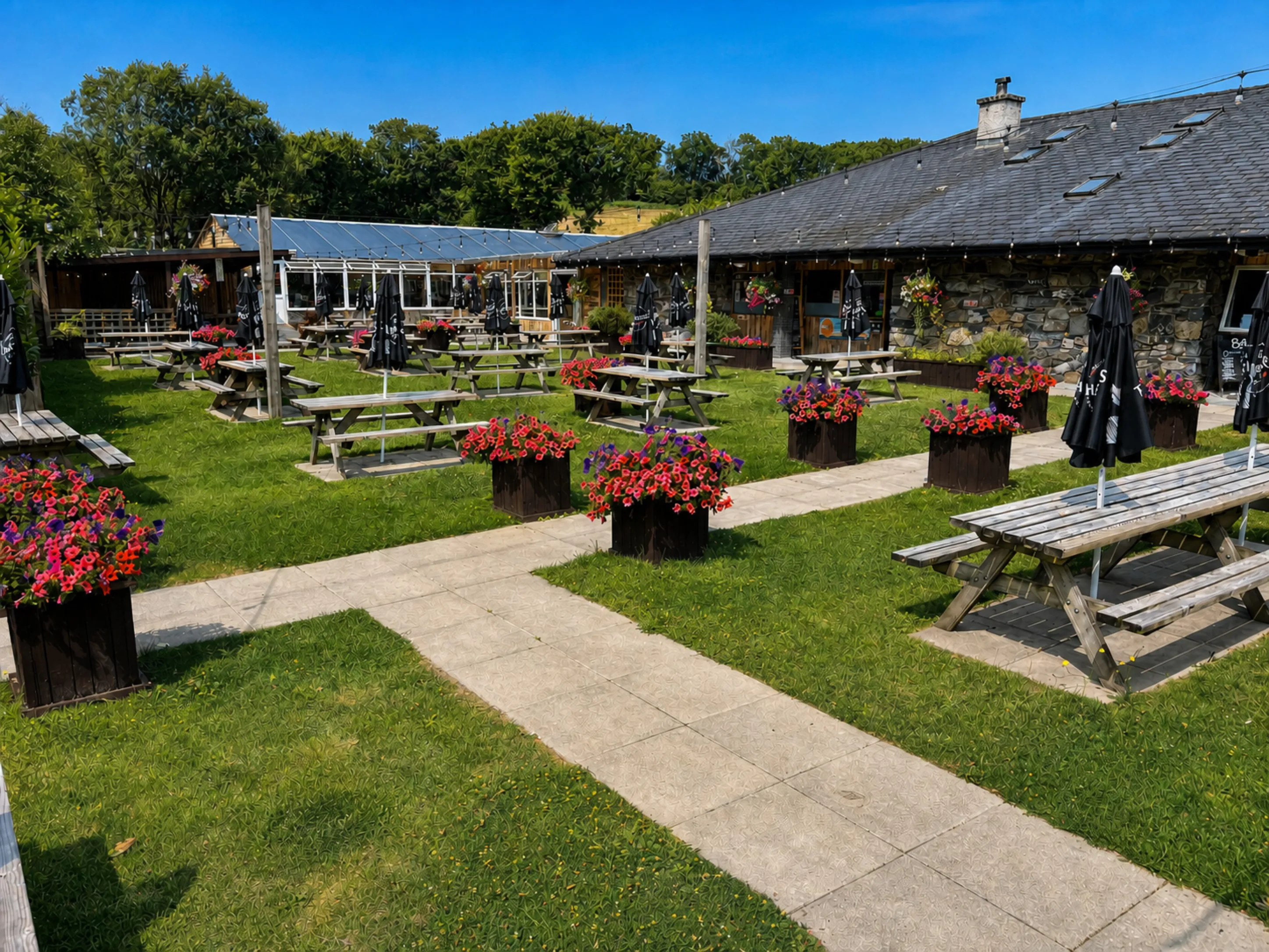 The beer garden in summer: picnic tables and umbrellas on the lawn, the stone pub behind, hills beyond