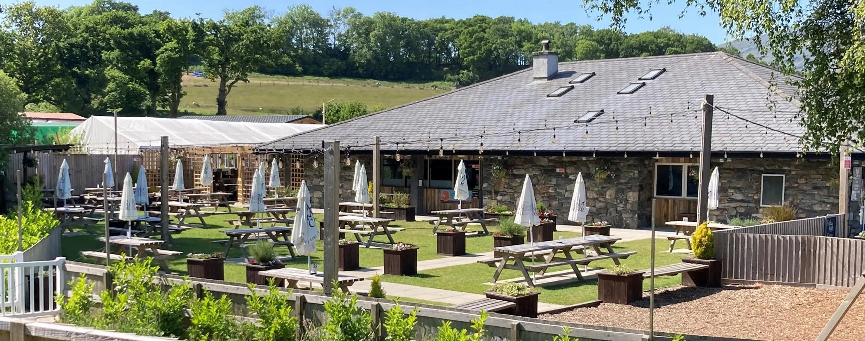 The Bryn Arms from the garden: the long stone building under a slate roof, picnic benches and parasols on the lawn, wooded hillside behind