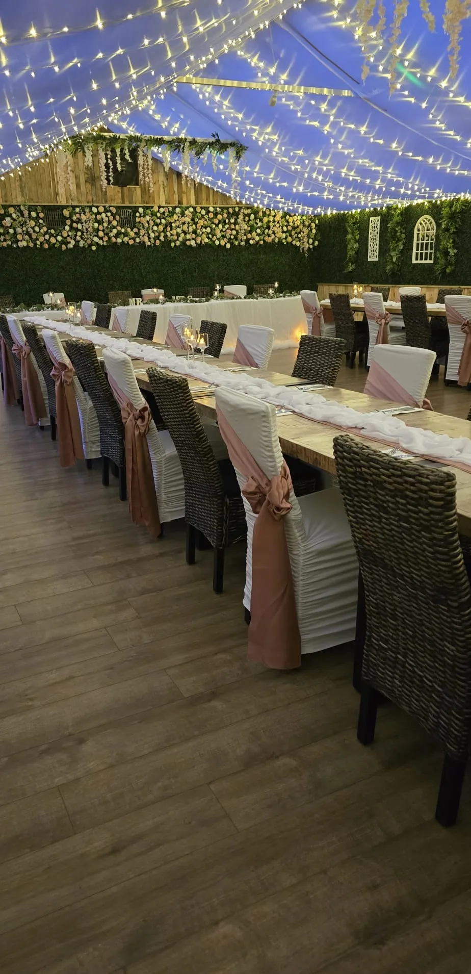 Long tables at dusk with blush sashes, a flower wall and a fairy light canopy